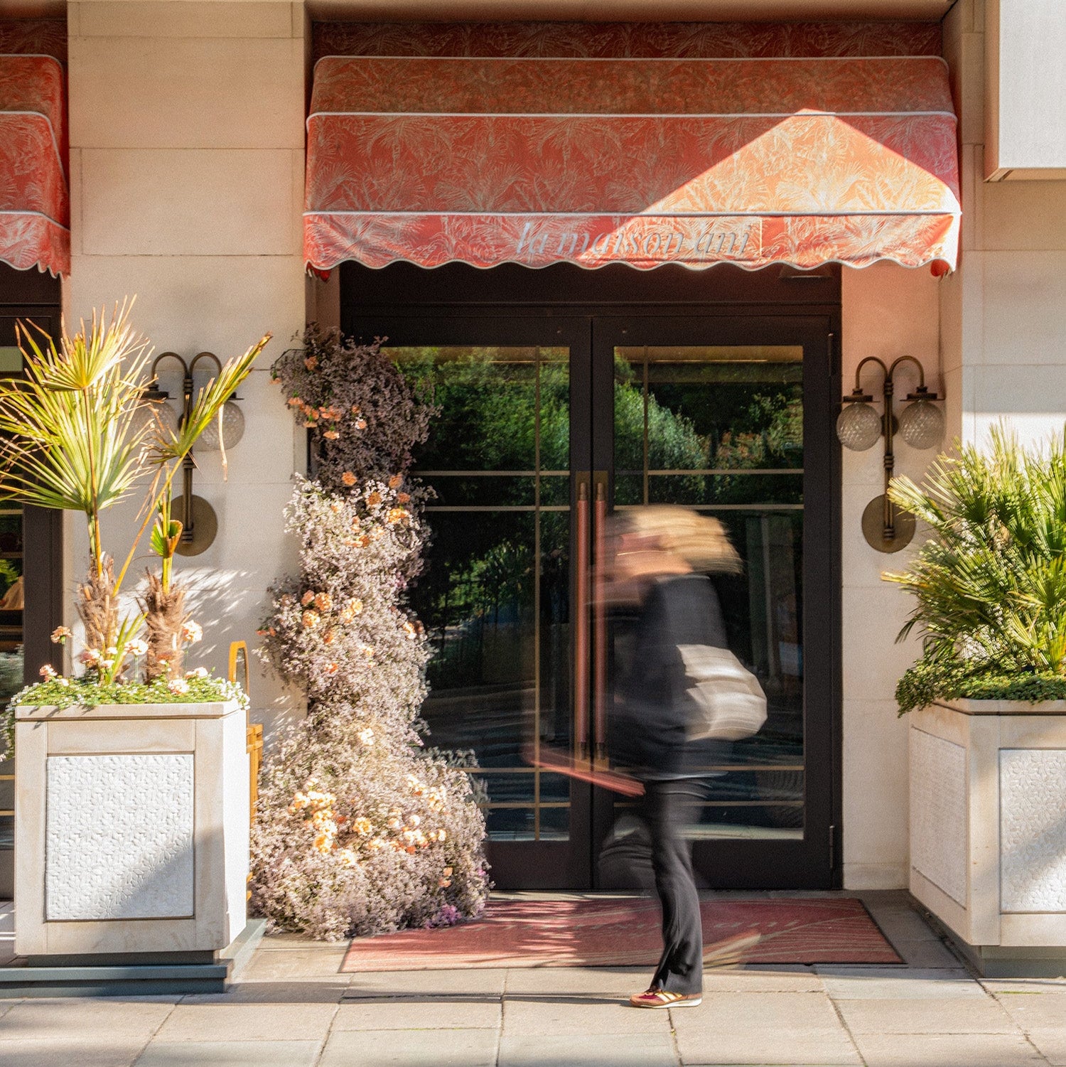 Person walking past a store entrance with decorative plants and an awning.