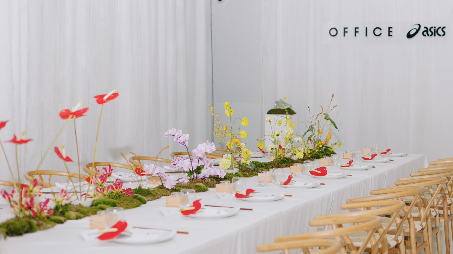 Decorated table with flowers and white plates against a white curtain backdrop, featuring Asics logo.