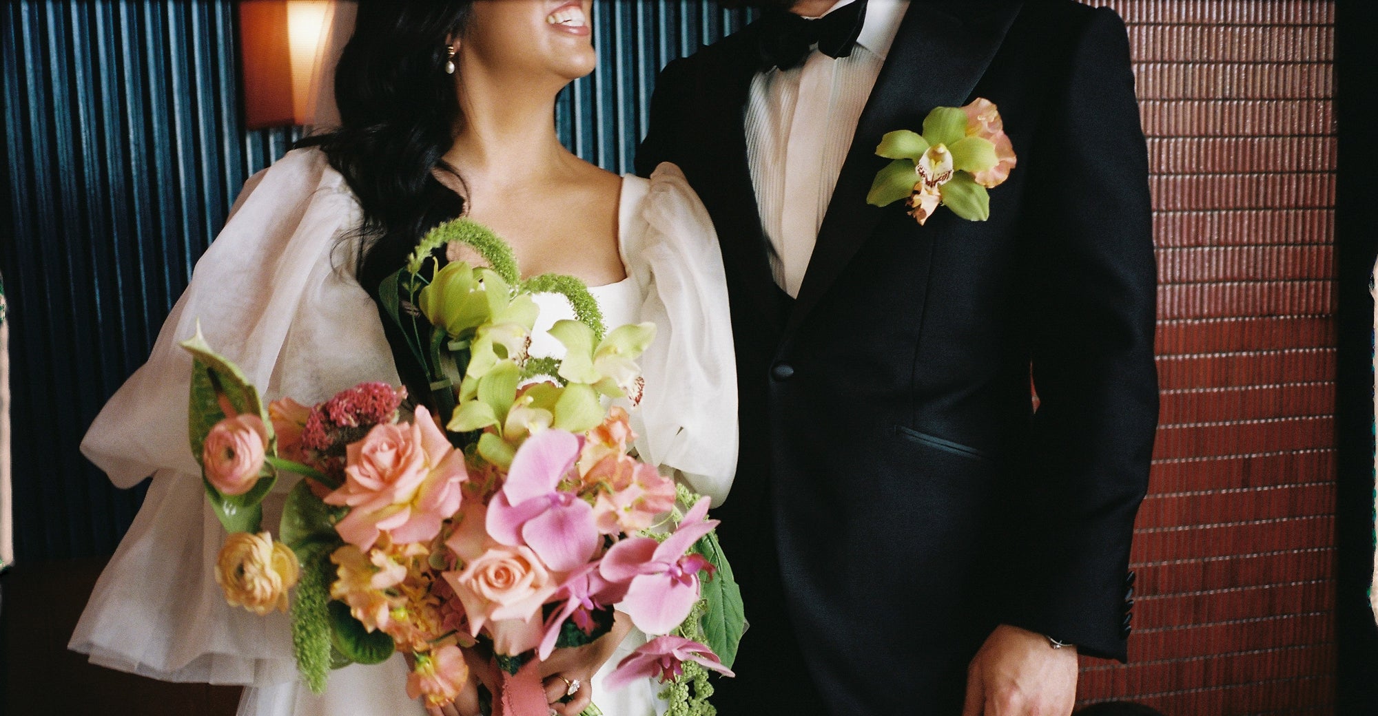 Man in a black suit with a floral boutonniere and woman in a white dress holding a bouquet, standing close together.