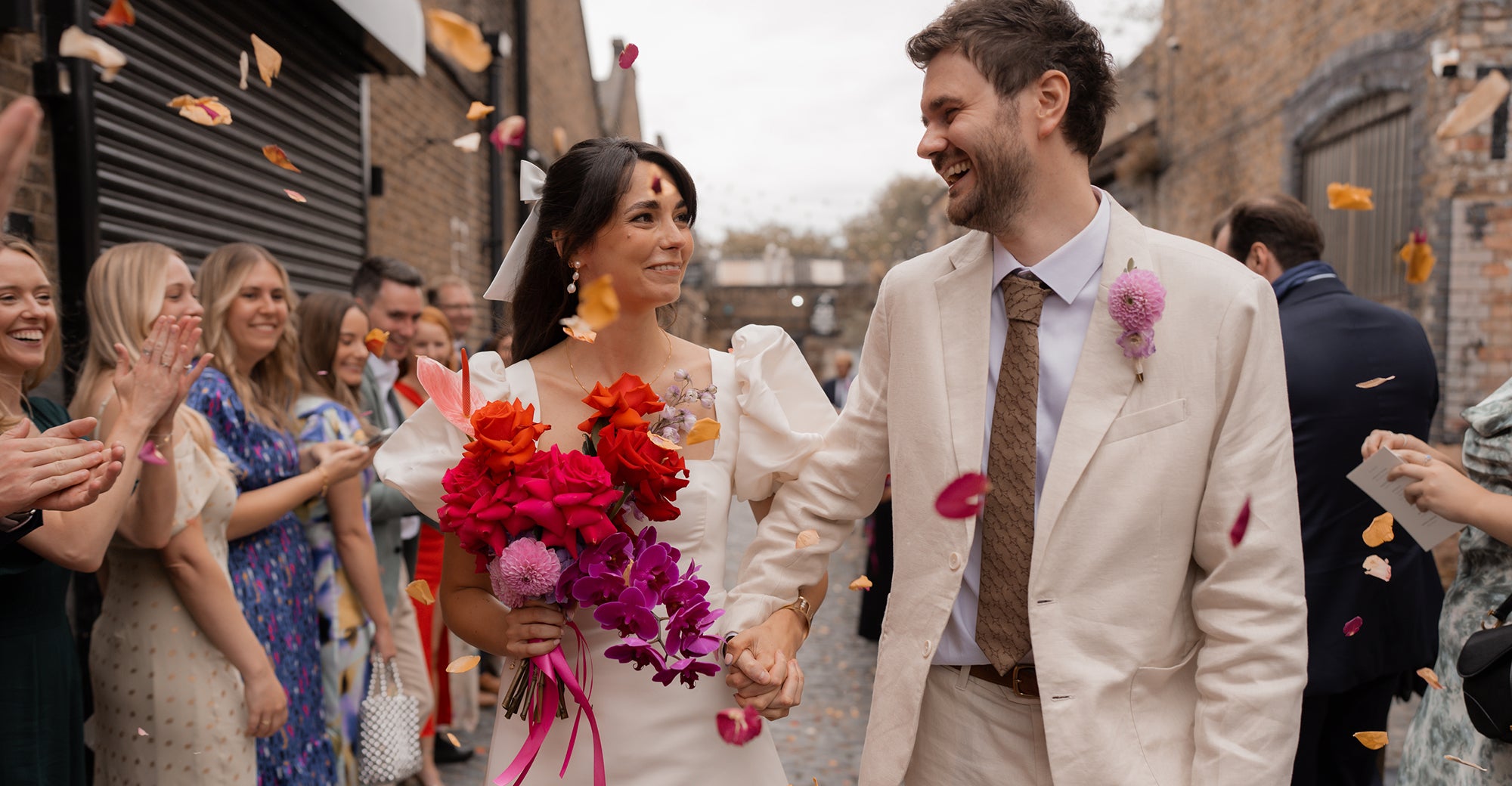 Wedding couple walking down a street with guests clapping, holding hands, and flowers being thrown.