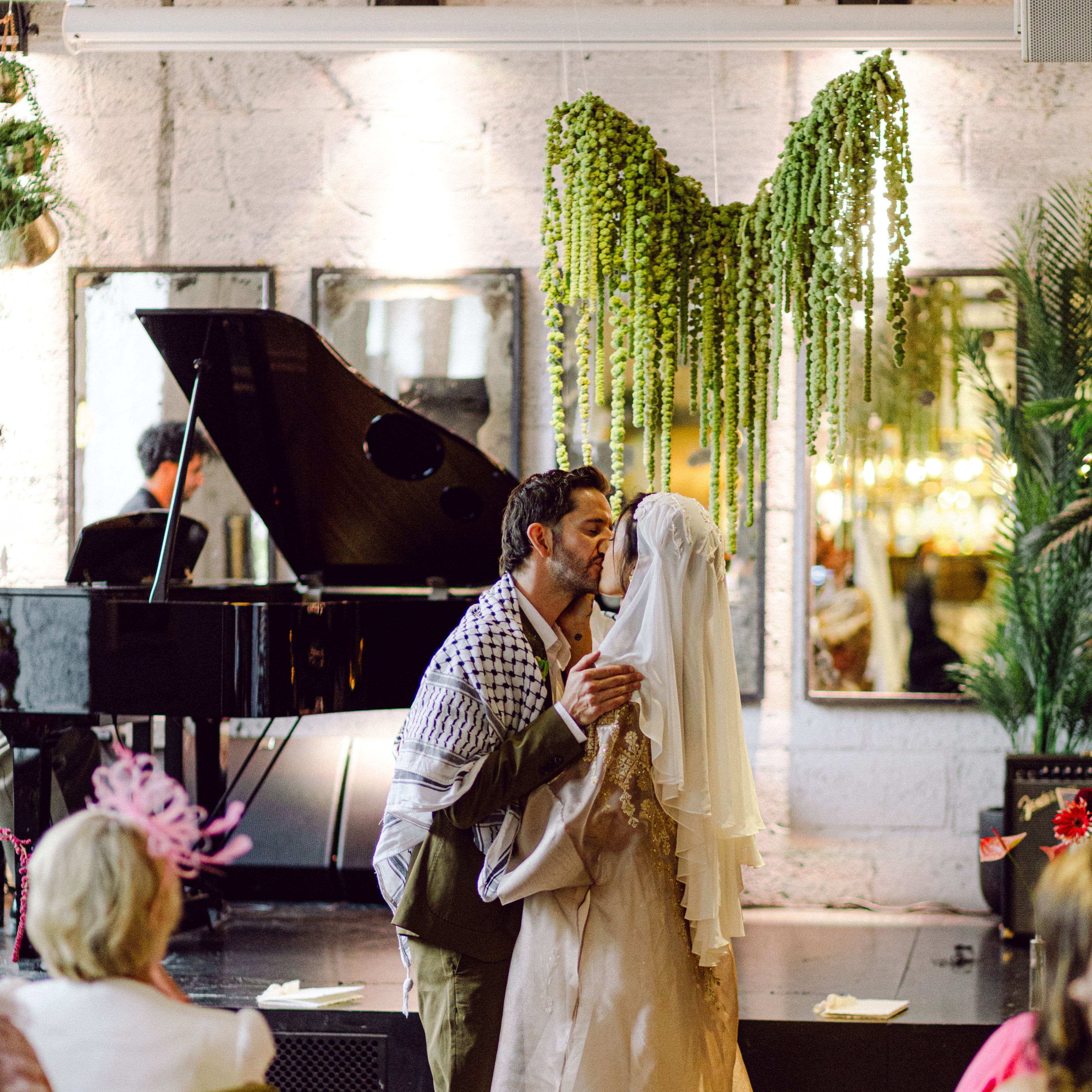 Couple kissing in front of a grand piano with plants and people in the background